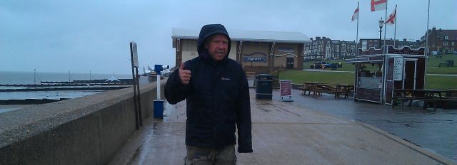 a man wearing a big rain coat looking very cold and wet on the sea front, giving the thumbs up